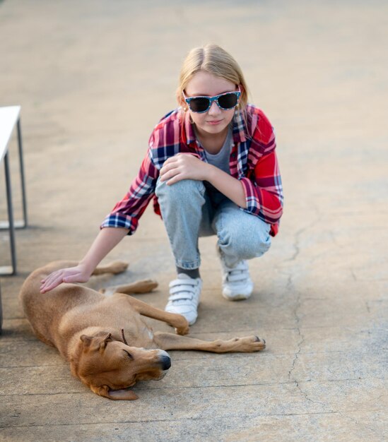 A teenage girl in a plaid shirt and sunglasses strokes a sleeping dog under a bench in a city park on a sunny summer day