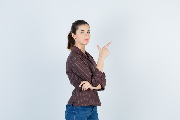 Teenage girl pointing up, standing sideways in brown striped shirt and looking serious. .