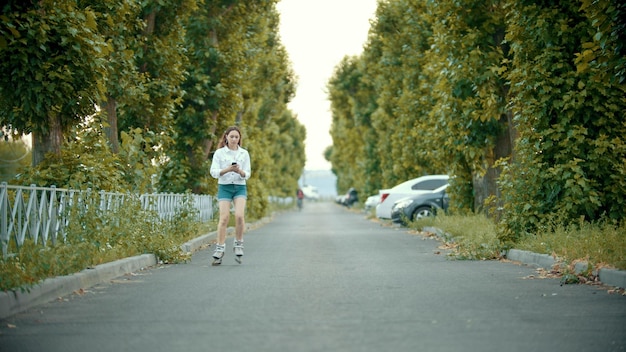 A teenage girl in rollerblades skating on the road looking at the phone