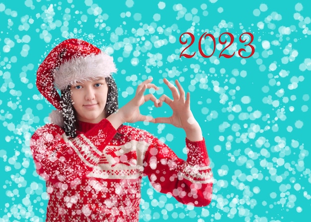 A teenage girl in a Santa Claus hat and a red Christmas sweater holds her hands in the form of a heart The concept of Christmas and New Year