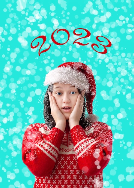 Photo a teenage girl in a santa claus hat and a red christmas sweater is surprised and holds her hands on her cheeks the concept of christmas and new year