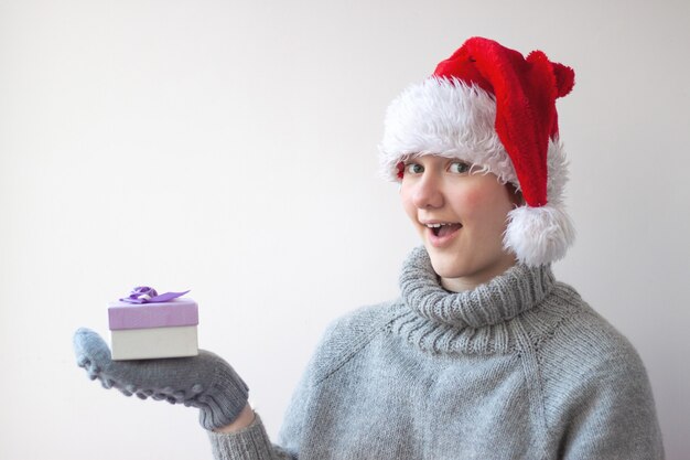 A teenage girl in a Santa hat and a gray knitted sweater and gray knitted mittens holds a small gift box in her hand and looks at the camera with surprise and joy