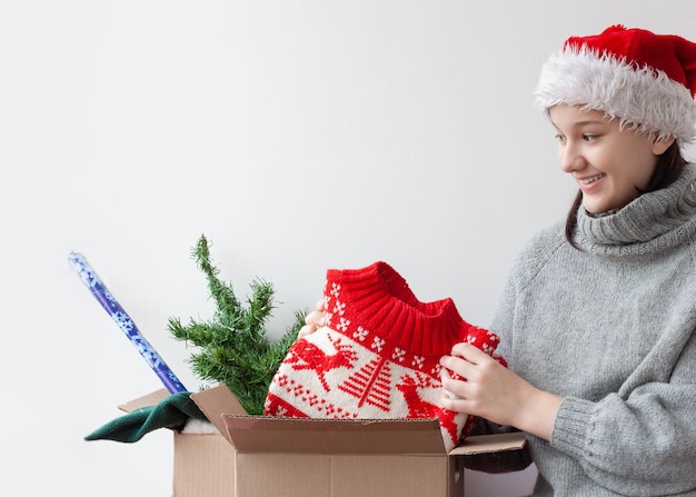 A teenage girl in a Santa hat takes out a Christmas sweater from a cardboard box