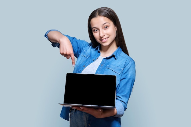 Teenage girl showing blank computer laptop screen posing against gray color studio background Teenager high school student pointing with her hand at space for copyspace text