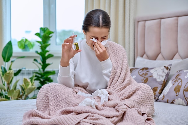 Teenage girl sitting in bed with runny nose holding medicine spray sneezing in tissue