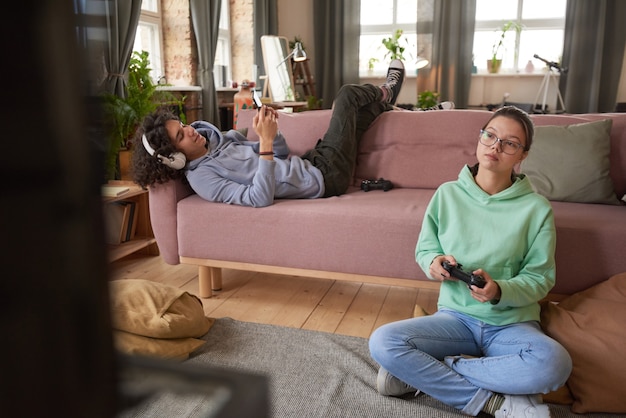 Photo teenage girl sitting on the floor playing video game with boy lying on sofa playing on the phone in the room