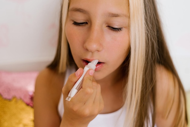 Teenage girl sitting at home on a bed with a cosmetic mirror paints her lips with lip gloss