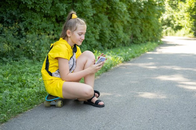 девочка-подросток сидит на скейтборде в парке с помощью мобильного телефона