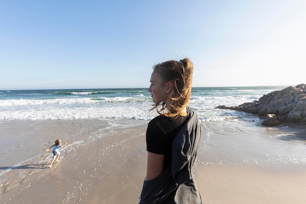 Teenage girl standing looking out over a beach a boy running on the sand below