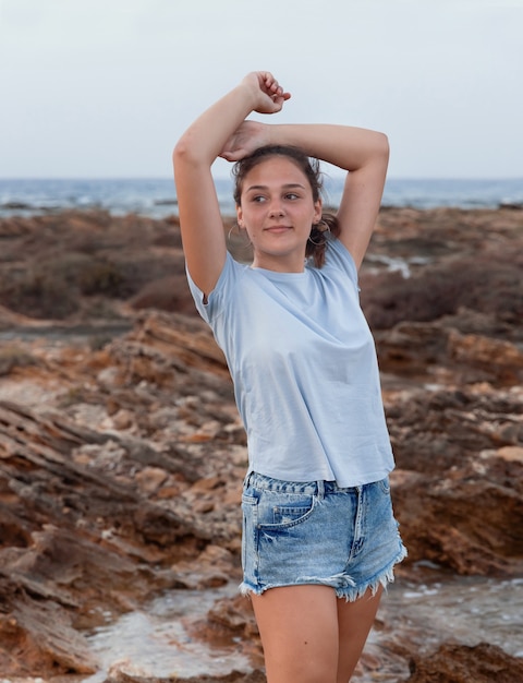 夕暮れ時の海沿いの崖の上に頭を抱えて立っている10代の少女は、水色のTシャツ、ジーンズのショートパンツを着ています。 4分の3の長さのショット。 Tシャツのモックアップ