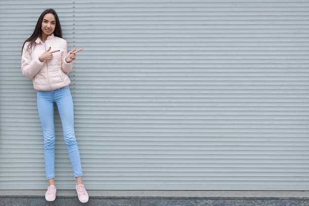 A teenage girl stands near a gray wall and points at it