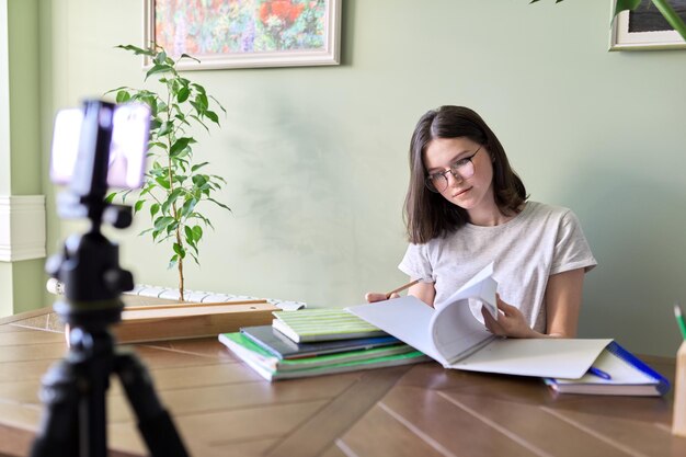 Teenage girl studies at home, sits at a table with books and notebooks using a smartphone, video call conference remote lesson. Technologies in education, online learning