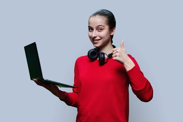 Teenage girl using laptop showing thumbs up gesture ok on grey background