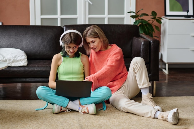 Teenage girl using laptop together with her mother while they sitting on the floor in living room