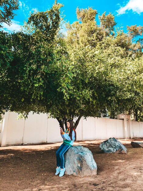 Teenage girl using mobile phone while sitting on rock against tree