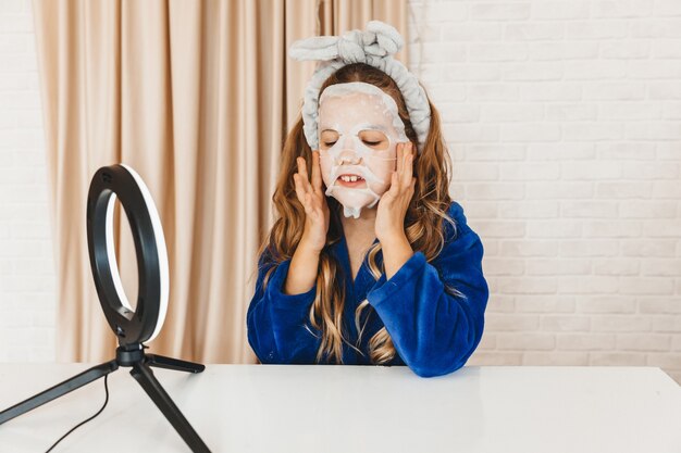 Teenage girl vlogger applying skin care face mask while recording video in front of light lamp. Happy girl having fun on filming tutorial using mobile smart phone.