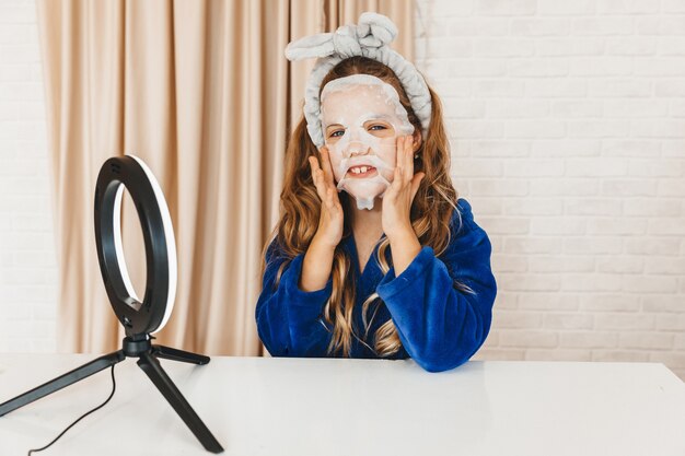 Teenage girl vlogger applying skin care face mask while recording video in front of light lamp. Happy girl having fun on filming tutorial using mobile smart phone.