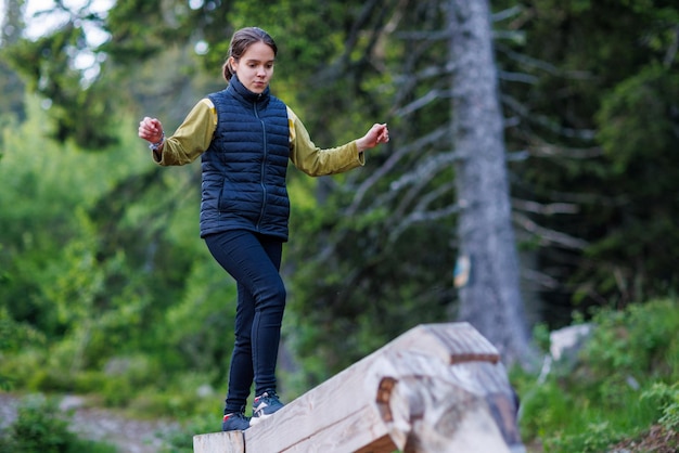 Teenage girl walks along an inclined log and passes a forest obstacle course in a pine forest