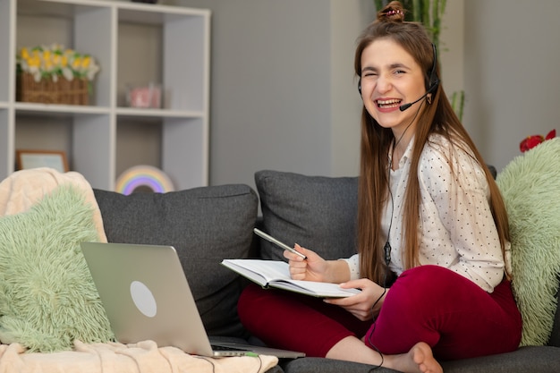 Teenage girl wearing headphones using laptop sitting on bed. Happy teen school student conference calling on computer for online distance learning communicating with friend by webcam at home