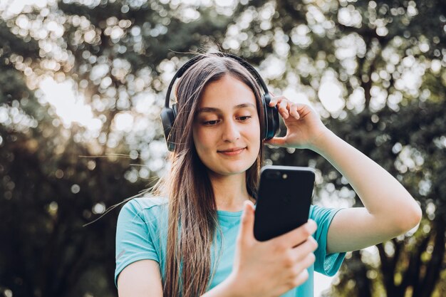 ターコイズのTシャツを着た10代の女の子、公園のスマートフォンでヘッドフォン設定プレイリストを使用