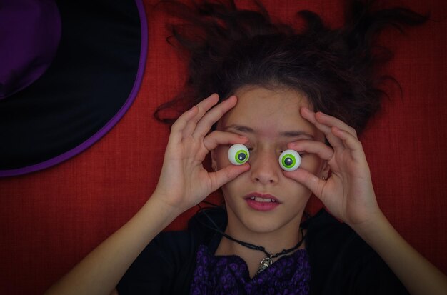 A teenage girl in a witch costume closes her eyes with human eyeshaped candy lying on an orangered couch with a halloween hat closeup top view