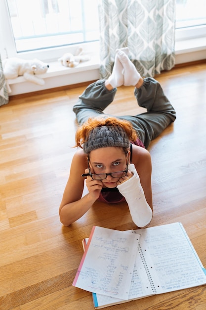 Девочка-подросток со сломанной рукой в гипсе в очках делает домашнее задание дома