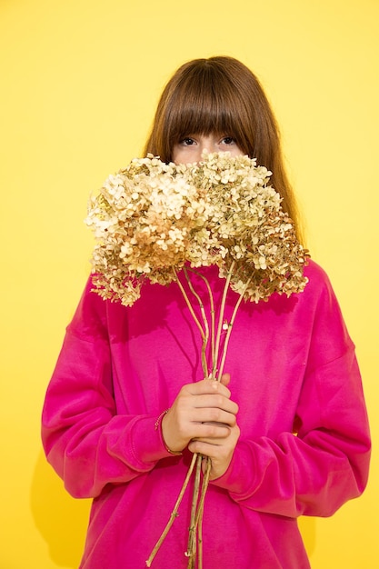 검은 머리와 앞머리를 가진 10대 소녀가 꽃 뒤에 숨었습니다. 분홍색 스웨터를 입고 노란색 배경의 스튜디오 사진, 초상화