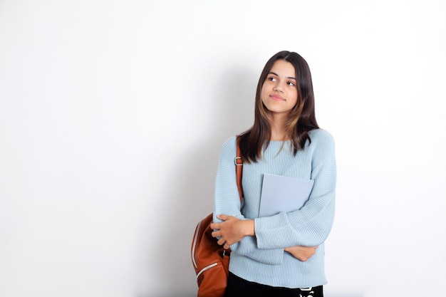 A teenage girl with dark hair with a backpack and a notebook on
a white background the schoolgirl thought about it student back to
school place for text