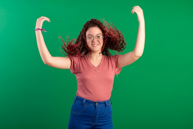안경을 쓴 10대 소녀가 자르기에 이상적인 녹색 배경으로 촬영된 스튜디오에서 곱슬곱슬한 빨간 머리를 재미있는 포즈로 흔들고 있습니다.
