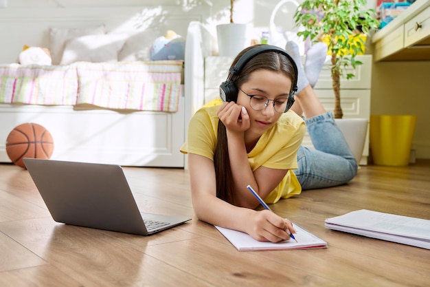 写真 眼鏡をかぶった10代の女の子がノートパソコンを使って自宅で勉強していますヘッドフォンと教科書をかけて部屋の床に横たわっている女性教育青春高校のコンセプト