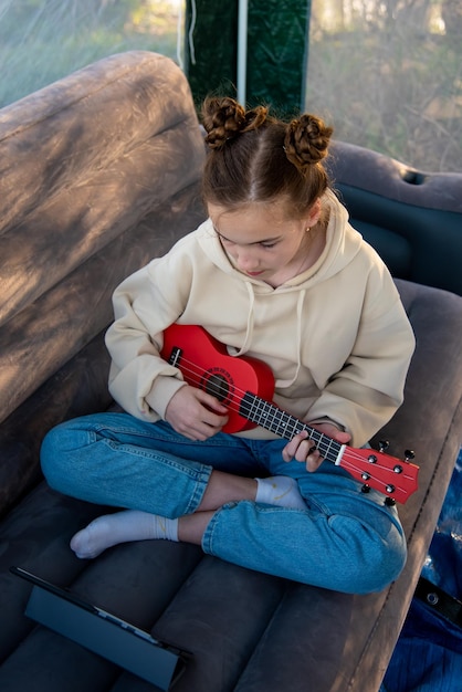 A teenage girl with a little guitar ukulele communicates online on the gadget.