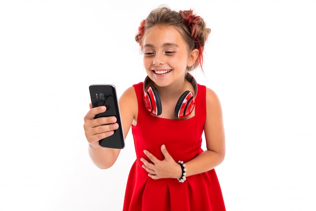 Teenage girl with long blonde hair, dyed tips pink, stuffed in two tufts, in red dress, with red headphones, bracelet, standing and holding phone in hand and laughing