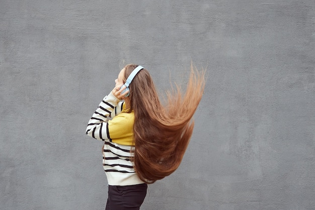 Фото Девочка-подросток с длинными волосами, развевающимися на ветру, слушает музыку в наушниках на сером фоне