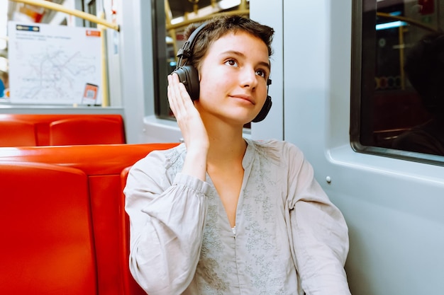 teenage girl with short haircut, smiling, listening to music in headphones while sitting in subway