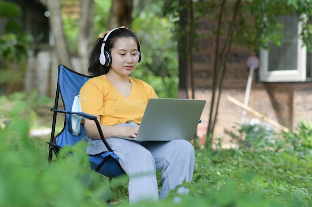 Ragazza adolescente che lavora su un laptop nel giardino di casa con le cuffie seduto su una sedia da campeggio