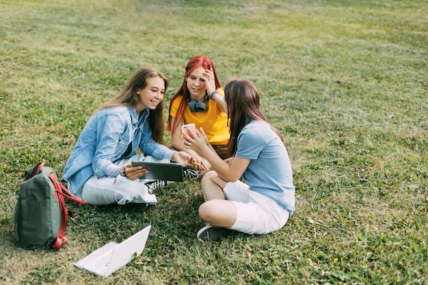 Photo teenage girls are sitting on a green lawn in a park with a backpack and a digital tablet