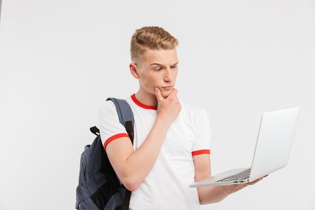 teenage guy 16-18 years old in t-shirt with backpack holding open laptop and seriously looking at screen isolated on white