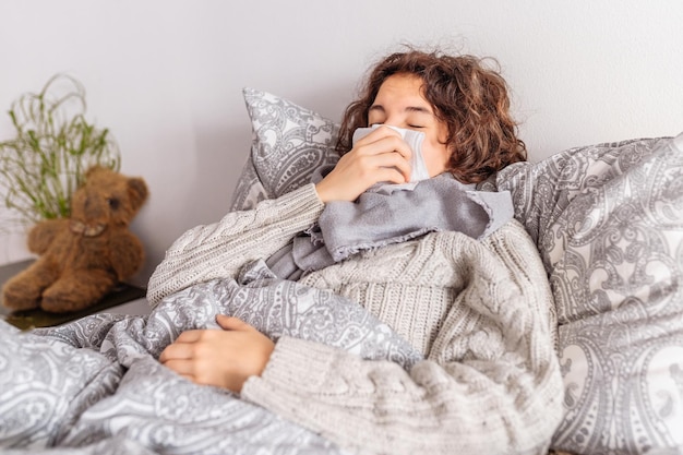 teenage lying in bed with warm scarf wrapped around throat in knitted sweater sneezes covering