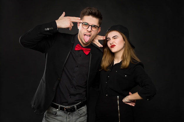 Подростковая улыбающаяся рэп-пара в черной одежде и черной кепке показывает знаки руками и языком. Изолированные цвета на черном фоне.