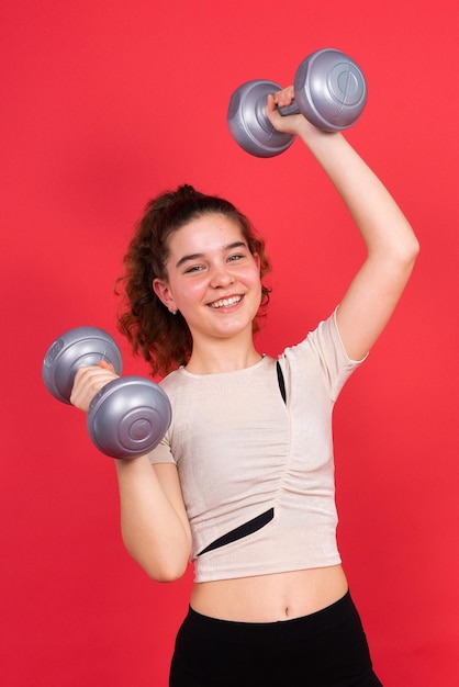 Teenage sportive girl exercises with dumbbells to develop muscles isolated on red background