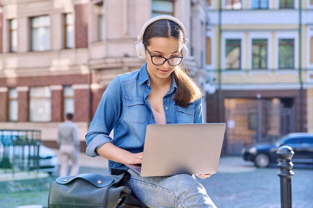 Teenage student girl in headphones using laptop outdoor urban background