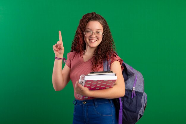 Teenage student wearing glasses wearing a school bag and carrying notebooks poses in a studio photo with a green background ideal for cropping