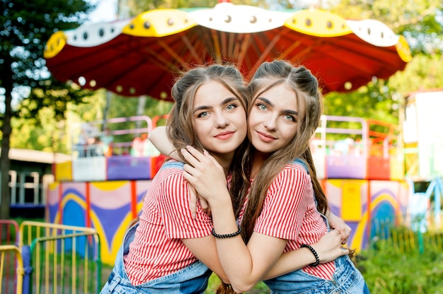 Sorelle gemelle adolescenti in abiti colorati che abbracciano, emozioni felici, con altalene colorate