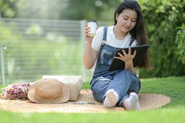 Una donna adolescente si siede nell'erba rilassante con un tablet e una tazza di caffè in chat online