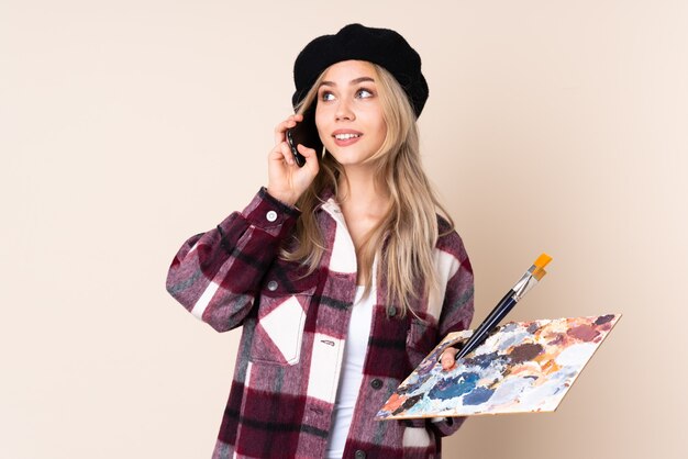 Teenager artist girl holding a palette on blue wall holding coffee to take away and a mobile