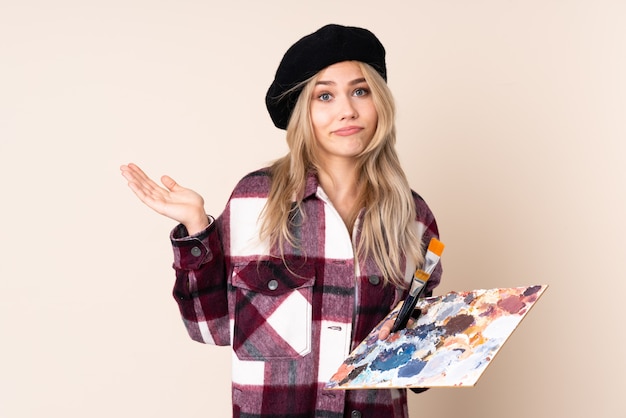 Teenager artist girl holding a palette on blue wall making doubts gesture