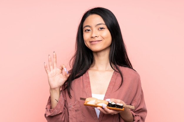 Ragazza asiatica dell'adolescente che mangia i sushi isolati sulla parete rosa che mostra un segno giusto con le dita