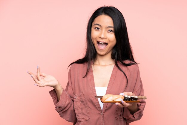 Teenager Asian girl eating sushi on pink wall unhappy and frustrated with something