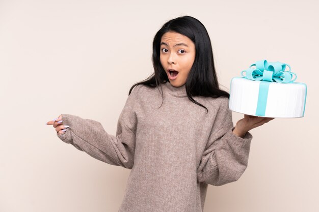 Teenager Asian girl holding a big cake isolated on beige background surprised and pointing side