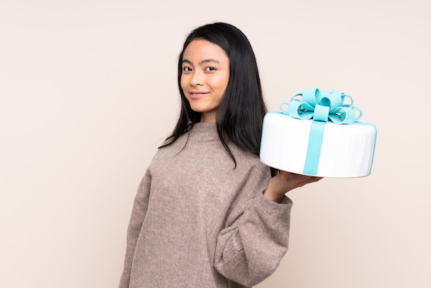 Teenager Asian girl holding a big cake isolated on beige smiling